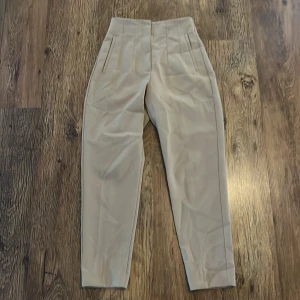 Beige kostymbyxor - Snygga beige kostymbyxor i mycket bra skick. De har en stilren design med hög midja och raka ben. Perfekta för både kontoret och festligare tillfällen. Materialet känns mjukt och bekvämt, vilket gör dem sköna att bära hela dagen. Obs! Har lite tråd i midjan som är lös men borde gå och klippa bort🤗