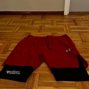 Dubbel träningsshorts från Under Armour - Säljer ett par röda träningsshorts från Under Armour i mycket bra skick. De har en elastisk midja med dragsko och svarta detaljer längst ner. Perfekta för träning eller en aktiv dag. Fickor finns på bägge sidorna, under låret och på baksidan. Passar S! Endast: 249