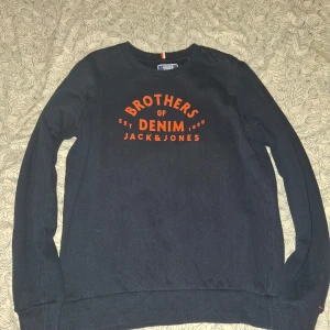 Mörkblå tröja från Jack & Jones - Säljer en mörkblå tröja från Jack & Jones med orange text 'Brothers of Denim' på framsidan. Tröjan har långa ärmar och är i mycket bra skick. Perfekt för en casual look eller en chill dag hemma. Passar bra till jeans eller chinos!