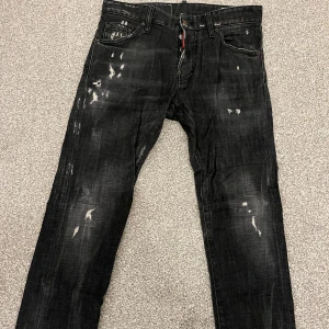 Äkta Dsquared2 Svarta jeans med slitningar - Säljer ett par svarta jeans från  Dsquared2 nästan nyskick. Notera ena knappen på gylfen utbytt, ej original. De har coola slitningar och en straight passform. Perfekta för en avslappnad stil med en edgy touch. Passar både till vardags och fest! Köpt för 4800 kr