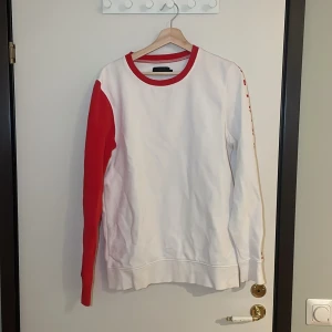 Vit och röd tröja från Produkt - Säljer en snygg vit tröja med röda detaljer från Produkt. Den har långa ärmar med text längs ena ärmen och en röd krage. Perfekt för en casual look eller en chill dag hemma. Passar bra till jeans eller joggers! Pris går alltid att förhandlas!