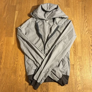 Grå vindjacka med huva - Säljer en grå vindjacka med huva från Standard American, perfekt för blåsiga dagar. Jackan har dragkedja framtill och muddar vid ärmslut och nederkant för extra komfort. Den är lätt och smidig att bära med sig. Passar bra till både vår och höst.