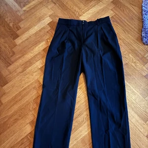 Svarta kostymbyxor - Säljer ett par stilrena svarta kostymbyxor i mycket bra skick. De har en hög midja och en rak passform som ger en klassisk look. Perfekta för både kontoret och festligare tillfällen. Byxorna har en dragkedja framtill och är gjorda i ett bekvämt material.
