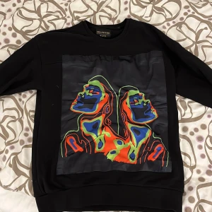Svart sweatshirt med färgglatt tryck - Säljer en svart sweatshirt från Bravado med ett coolt, färgglatt tryck på framsidan. Perfekt för att liva upp garderoben! Tröjan har långa ärmar och är i bra skick. Passar både till vardags och festliga tillfällen. Passar M o L