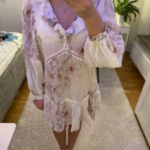 Blommig vit klänning - Säljer en superfin blommig klänning i vitt med volanger och spetsdetaljer. Den har långa ärmar och ett romantiskt mönster i ljusa toner. Perfekt för vår och sommar! 🌸