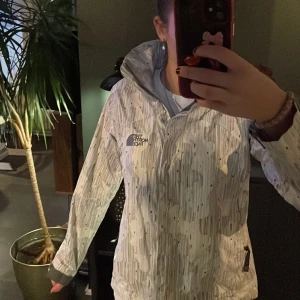 Vit vindjacka från The North Face - Säljer en snygg vit vindjacka från The North Face med ett diskret randigt mönster. Jackan har en dragkedja framtill och är perfekt för blåsiga dagar. Den har en normal passform och är långärmad. Perfekt för vår och höst!
