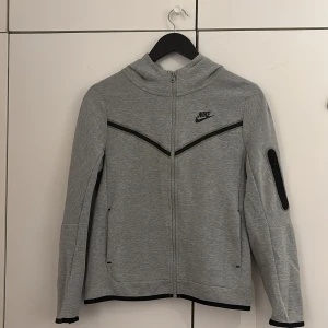 Grå huvtröja Nike Tech - Säljer en grå huvtröja från Nike i mycket bra skick. Den har en dragkedja framtill och svarta detaljer längs ärmarna och fickorna. Perfekt för höst och vår, och passar bra till träning eller vardagsbruk. Superbekväm och stilren design!