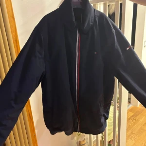 Svart jacka från Tommy Hilfiger - Säljer en snygg svart bomberjacka från Tommy Hilfiger. Den har en klassisk design med dragkedja i rött, vitt och blått. Jackan är långärmad och perfekt för höst och vår. Den är i mycket bra skick och passar både till vardags och fest.