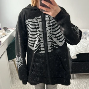 Svart hoodie med skelettmönster - Cool svart hoodie med ett vitt skelettmönster på framsidan och spindelnätsdetaljer på ärmarna. Perfekt för höst och Halloween! Den har en dragkedja framtill och är i mycket bra skick. Passar perfekt för en avslappnad och edgy stil. Den är från TEMU och är over sized xs