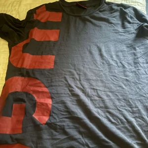 Svart t-shirt från Hugo - Säljer en svart t-shirt från Hugo med stora röda bokstäver på framsidan. Den är kortärmad och perfekt för en casual look. T-shirten är i bra skick och passar både till vardags och fest. Skön och stilren design!