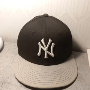 Svart och grå New Era keps - Säljer en snygg svart och grå keps från New Era, modell 9FIFTY. Den har en broderad logga framtill och justerbar passform baktill. Perfekt för både vardag och sportiga aktiviteter. Kepsen är i okej skick och redo för nya äventyr!