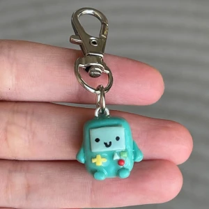 Turkos nyckelring med spelkonsolfigur - Söt turkos nyckelring med en liten spelkonsolfigur. Den har ett glatt ansikte och detaljer i gult och rött. Perfekt att fästa på väskan eller nycklarna för en personlig touch. Nyckelringen är cirka 4 cm lång inklusive metallklämman.