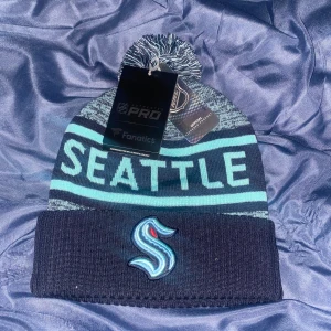 Seattle Kraken mössa från Fanatics - Snygg och varm mössa från Fanatics med Seattle Kraken-tema. Den är mörkblå med turkosa detaljer och texten 'SEATTLE' och 'KRAKEN' i stora bokstäver. Mössan har en tofs på toppen och ett broderat emblem framtill. Perfekt för kalla dagar och att visa ditt stöd för laget! Helt oanvänd och my med taggar kvar. Inköpt från USA för 35 dollar eller cirka 350kr. 