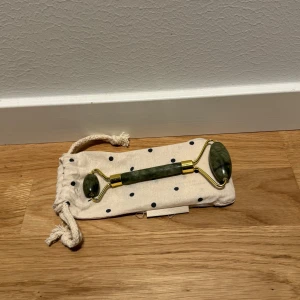 Grön ansiktsroller med gulddetaljer - Säljer en snygg ansiktsroller i grön sten med gulddetaljer. Perfekt för att massera ansiktet och ge huden en fräsch känsla. Kommer med en prickig tygpåse för förvaring. Perfekt för en avkopplande stund hemma!