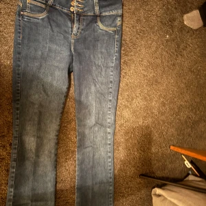 Blå jeans från JLO - Säljer ett par snygga blå jeans från JLO. De har en klassisk femficksdesign och är i bra skick. Jeansen har en normal passform och är perfekta för vardagsbruk. De är tillverkade i ett slitstarkt denimtyg och har en knappgylf framtill.