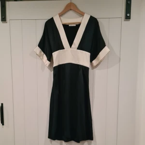 Svart och vit klänning HM Madonna - Elegant svart klänning med benvita detaljer och knytband i midjan. Från HM Madonna kollektion. Klänningen är i satinliknande tyg.