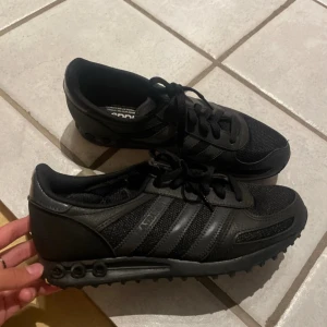 Svarta sneakers från Adidas - Säljer ett par svarta La Trainer sneakers från Adidas i nyskick. De har en stilren design med snörning och är perfekta för både vardag och träning. Skorna är gjorda i syntetmaterial och har en bekväm passform. Perfekta för höst och vår! 