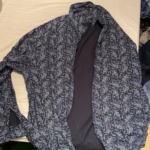 Mönstrad skjorta från Dressmann - Snygg skjorta från Dressmann i ett stilrent paisleymönster. Skjortan är blå med vita detaljer och har en klassisk krage. Tillverkad i bomull med stretch för extra komfort. Perfekt för en trendig look.