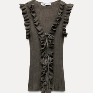 Olivgrön ribbad topp från Zara - Säljer en olivgrön ribbad topp från Zara med volangdetaljer längs ärmarna och framsidan. Toppen har en v-ringad design och knytband framtill. Perfekt för en stilren look.