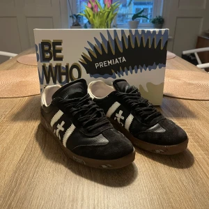  Premiata sneakers  - Snygga svarta sneakers från Premiata med vita detaljer och snörning. Skorna har en stilren design med en vit logotyp på sidan och en brun sula. Perfekta för en avslappnad stil.