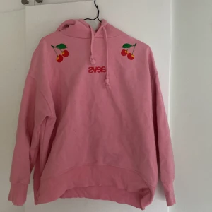 Rosa hoodie med körsbär från Svea - Säljer en söt rosa hoodie från Svea med körsbärsdetaljer. Hoodien har en justerbar huva och långa ärmar. Perfekt för en avslappnad stil.