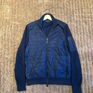 Mörkblå jacka från J.Lindeberg - Säljer en snygg mörkblå jacka från J.Lindeberg. Jackan har en quiltad framsida med dragkedja och stickade ärmar. Perfekt för kyligare dagar med sin stilrena design och bekväma passform.