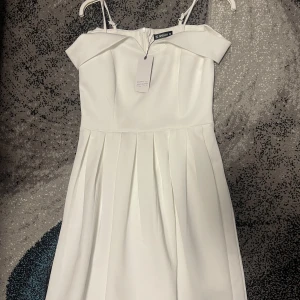 Vit klänning från Sheilay - Elegant vit klänning från Sheilay med offshoulder-design och smala axelband. Klänningen har en vacker veckad kjol och dragkedja i ryggen. Perfekt för speciella tillfällen och student. Prislappen på eftersom den aldrig har kommit till användning. Den är stor i storleken och sitter mer som M än S skulle jag säga. 