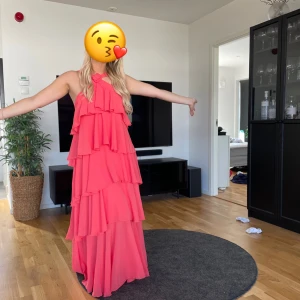 Koral balklänning  med volanger - Säljer en fantastisk rosa långklänning med flera lager av volanger. Klänningen har en öppen rygg och breda axelband som ger en elegant look. Perfekt för sommarens festligheter! 🌸