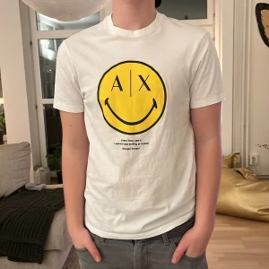 Vit t-shirt från Armani Exchange - Säljer en vit t-shirt från Armani Exchange med ett stort gult smileytryck och text på framsidan. T-shirten är kortärmad och har en normal passform. Perfekt för en avslappnad stil.