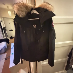 Svart parkajacka från Canada Goose Montebello  - Säljer en svart parkajacka från Canada Goose med pälsfodrad huva. Jackan har en ikonisk logga på ärmen och stängs med både dragkedja och knappar. Perfekt för kalla vinterdagar.