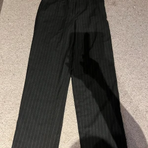 Svarta randiga byxor från H&M - Snygga svarta kostymbyxor från H&M med diskreta ränder. De har en klassisk straight passform. Byxorna är medelhöga i midja. Storlek 40. Priset kan ändras 