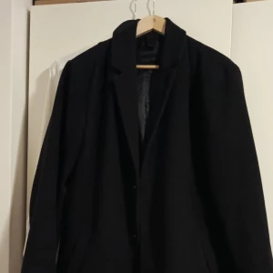 Svart kappa från KIOMI - Manlig, svart kappa från KIOMI i storlek L.  Har nyligen köpt en ny kappa och därför bestämt mig för att göra mig av med denna. Varsamt använd och är i gott skick!  Material: Ullblandning