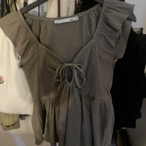 Brun volangtopp med knytning - Säljer en söt brun topp med volangdetaljer och knytning framtill. Toppen har en v-ringad design och korta ärmar. Perfekt för en avslappnad stil.