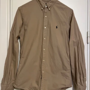 Beige/ljusbrun skjorta från Ralph Lauren - Snygg beige/ljusbrun skjorta från Ralph Lauren i slim fit. Skjortan har långa ärmar och knappar framtill samt en broderad logga på bröstet. Perfekt för en stilren look. Sparsamt använd i mycket gott skick. Se gärna mina andra annonser, så kan allt skickas i samma sändning. 