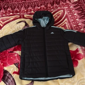Svart pufferjacka från Adidas - Säljer en svart pufferjacka från Adidas med huva och dragkedja. Jackan har långa ärmar och turkosa detaljer på axlarna. Perfekt för kyligare dagar. Har använt den hemma en gång bara för testa passformen men tyvärr var den för liten.