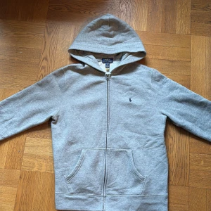 Grå hoodie från Polo Ralph Lauren - Säljer en stilren grå hoodie från Polo Ralph Lauren med dragkedja och huva. Tröjan har långa ärmar och en liten broderad logga på bröstet. Perfekt för en avslappnad look. 
