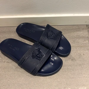 Mörkblå sandaler från Versace - Snygga mörkblå sandaler från Versace med det ikoniska Medusa-huvudet på remmen. Sandalerna har en stilren design med grekiskt mönster och är tillverkade i syntetmaterial. Perfekta för en avslappnad och trendig look. Skick 9,5/10 knappt använda.