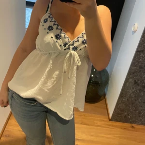 Vit topp med blåa fina broderier, gå in på profil för mer - Söt vit topp med blå blommiga broderier och knytning framtill. Den har en v-ringad design och smala axelband. Super fin unde tvären och sommaren. Är i storlek S men passar mig som bär XS