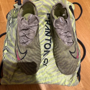 Nike Phantom fotbollsskor - Säljer ett par Nike Phantom fotbollsskor i grön/gul  med snörning. Skorna har en modern design med mönster och en slitstark sula för bra grepp på planen. Perfekta för fotbollsträning och matcher.