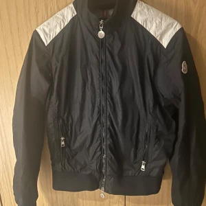 Svart vindjacka från Moncler - Säljer en vindjacka från Moncler med vita detaljer på axlarna. Säljer pga att jackan är för liten för mig men annars inga fel. Kom gärna med frågor👍