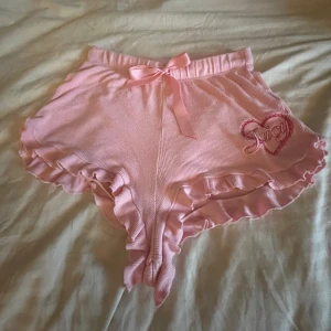Rosa shorts från Juicy Couture - Söta rosa shorts från Juicy Couture med volangdetaljer och en rosett framtill. Broderad hjärtdetalj med texten 'Juicy' på sidan. Perfekta för en avslappnad dag hemma. Ny pris 500kr