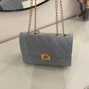 Ljusblå quiltad axelväska - Snygg ljusblå axelväska med quiltat mönster och guldfärgad kedja. Väskan har en elegant låsdetalj framtill och är perfekt för att ge en stilren touch till din outfit. Den kompakta storleken gör den smidig att bära med sig.