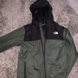 Grön och svart vindjacka från The North Face - Säljer en grön och svart vindjacka från The North Face. Jackan har en huva och dragkedja framtill. Den är långärmad med The North Face-logga på bröstet och ärmen. 