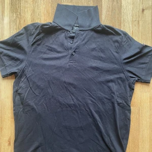 Svart pikétröja från Hugo Boss - Snygg svart pikétröja från Hugo Boss med klassisk krage och knappar framtill. Perfekt för en stilren look. Tröjan är kortärmad och har en diskret broderad logga på bröstet. Storlek: medium. Hör av dig vid några frågor.😊