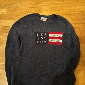 Mörkblå stickad tröja från Lexington - Säljer en snygg mörkblå stickad tröja från Lexington med text och mönster i rött och vitt på framsidan. Tröjan har långa ärmar och en klassisk rund halsringning. Perfekt för kyligare dagar! 