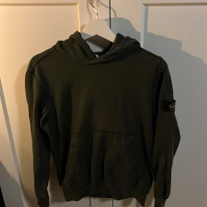 Grön hoodie från Stone Island Junior - Säljer en snygg grön hoodie från Stone Island Junior. Den har en klassisk design med huva och en praktisk magficka. På ärmen finns det ikoniska Stone Island-märket. Perfekt för en stilren och avslappnad look.