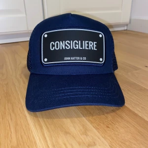 Marinblå keps från John Hatter & Co - Tjena! Säljer nu denna snygga marinblåa keps från John Hatter & Co med texten 'Consigliere' på framsidan. Kepsen har ett nätmaterial på baksidan för bättre ventilation och en justerbar rem för perfekt passform. Den är i nyskick och har bara testats ett fåtal gånger.  Nypris: 500 kr  Mitt pris: 199 kr (öppen för prisförslag)  Tveka inte på att höra av dig om du har några frågor eller vill se fler bilder! 