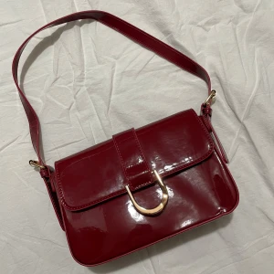 Röd axelväska med gulddetalj - Snygg röd axelväska i glansigt material med en elegant guldfärgad spänne framtill. Väskan har en justerbar axelrem och en stilren design som passar perfekt för att lyfta din outfit. Perfekt för att bära dina nödvändigheter med stil.
