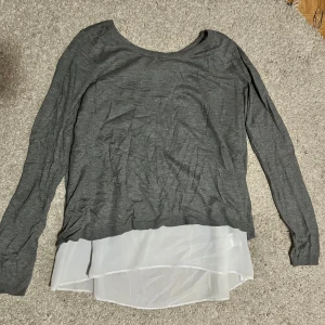 Grå tröja med vit detalj - Snygg grå tröja med lång ärm och vit chiffongdetalj som ger en lager-på-lager-effekt. Perfekt för en stilren look med en twist. Den har en öppen rygg som ger extra stilpoäng. Passar bra till både jeans och kjol.