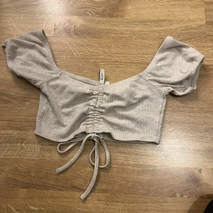 Beige ribbad topp från NLY Trend - Säljer en söt beige ribbad topp från NLY Trend. Toppen är croppad med korta ärmar och har en knytning framtill för en justerbar passform. Perfekt för varma dagar!